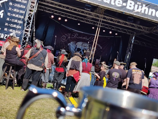 Einige Personen in Piratenkostümen vor einer Bühne. Im Bild Vordergrund der Rand eines Metallkruges. Das Banner im Hintergrund der Bühne kündigt die Band 