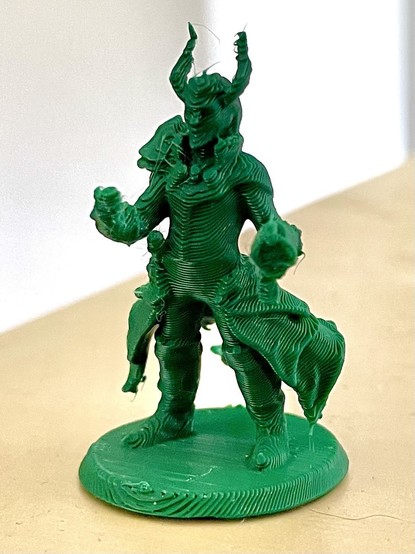 Eine grüne 3D-gedruckte Figur eines Charakters mit Hörnern und einem verzierten Outfit, die auf einer runden Basis steht.