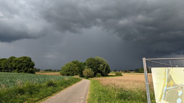Vorne ein Weg zwischen Feldern in der Sonne und im Hintergrund dunkle Wolken mit Regen