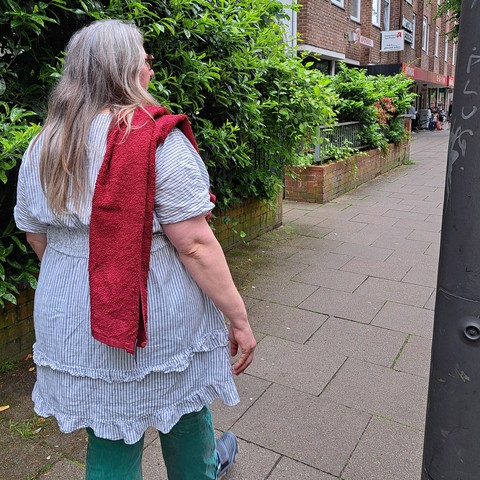 Frau mit grauen langen Haaren geht mit rotem Handtuch über der Schulter durch die Straße. Man sieht sie von hinten.