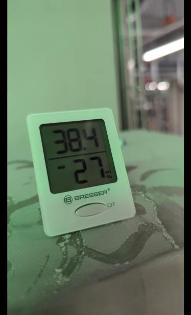 Kleines, weißes Haushaltsthermometer, das einen Wert von 38,4 Grad Celsius und eine Luftfeuchtigkeit von 27%