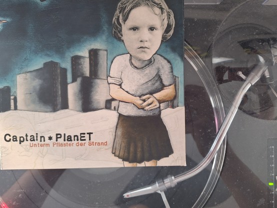 Das Cover der 7-inch-Vinyl-EP Unterm Pflaster der Strand von Captain planet auf dem laufenden Plattenspieler. Es zeigt im einer Mischung aus Zeichnung und Foto ein Kind, das einem Rock trägt, vor einer angedeuteten Stadt. Der Blick wirkt traurig oder desillusioniert. In der oberen Ecke fliegt ein Drachenflieger. 