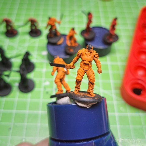 Nahaufnahme einer 2cm großen Spielfigur in Form eines Soldaten mit Schwert in Hand. Figur ist komplett Orange, Kopf schwarz. Figur klebt an einem Plastikstück zum besseren Halten. 
Im Hintergrund verschwommen weitere dieser Miniaturen.