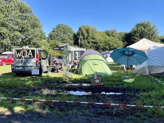 Campingszene mit Zelten, einem Lieferwagen, einem Regenschirm und einem abgeklebten schlammigen Bereich. Bäume und klarer blauer Himmel im Hintergrund.