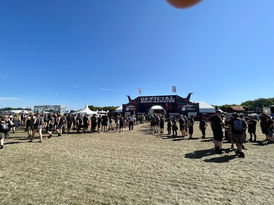 Leute, die am offiziellen Merchandise-Stand eines Outdoor-Festivals unter einem klaren blauen Himmel in der Schlange stehen.