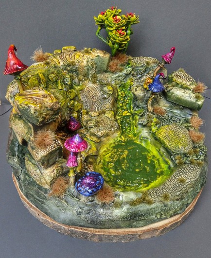 Photo of a little round diorama. It's a pool amidst some ruins with lots of water and mushrooms. The pool itself isn't filled yet and the miniatures are still missing.